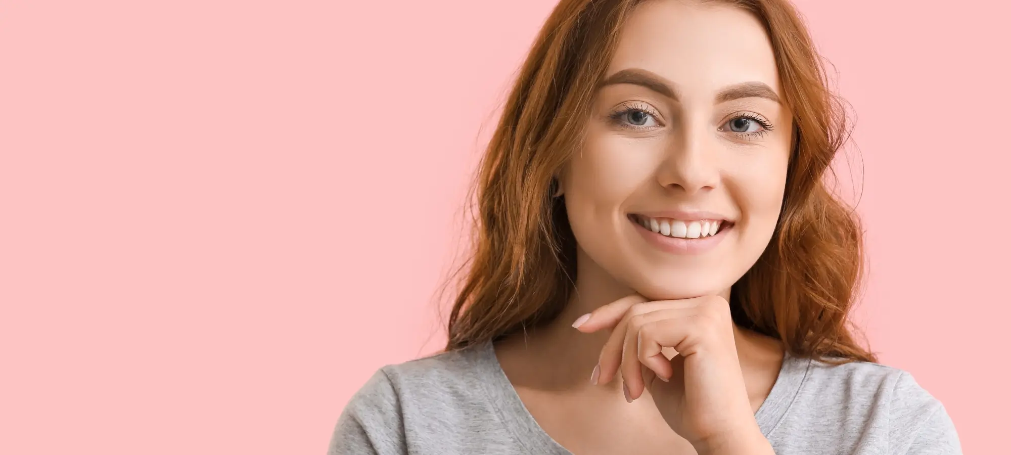 Young redhead resting her face on her relaxed hand on a light pink background.