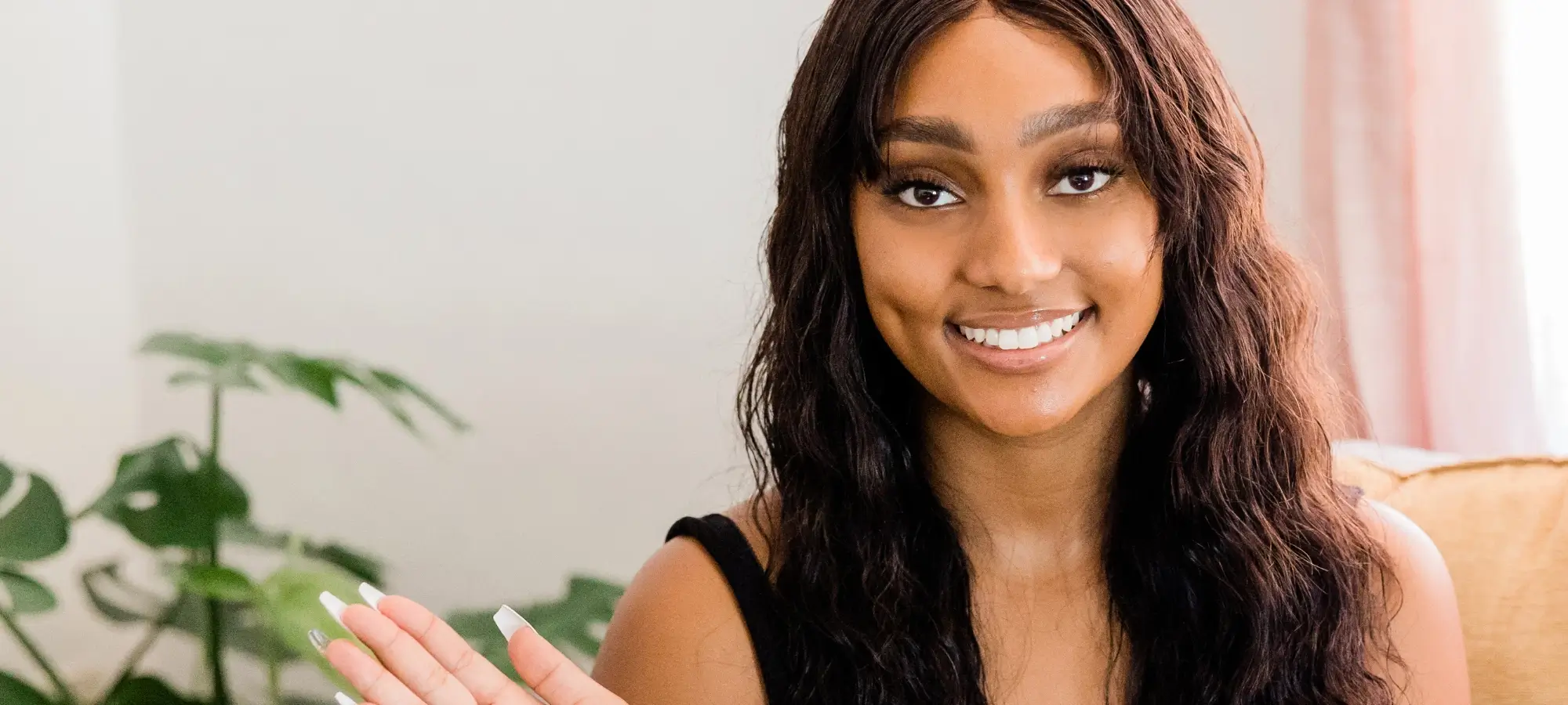 Young black woman with long hair smiling and waving a manicured hand, sitting on a plush chair in a comfortable environment