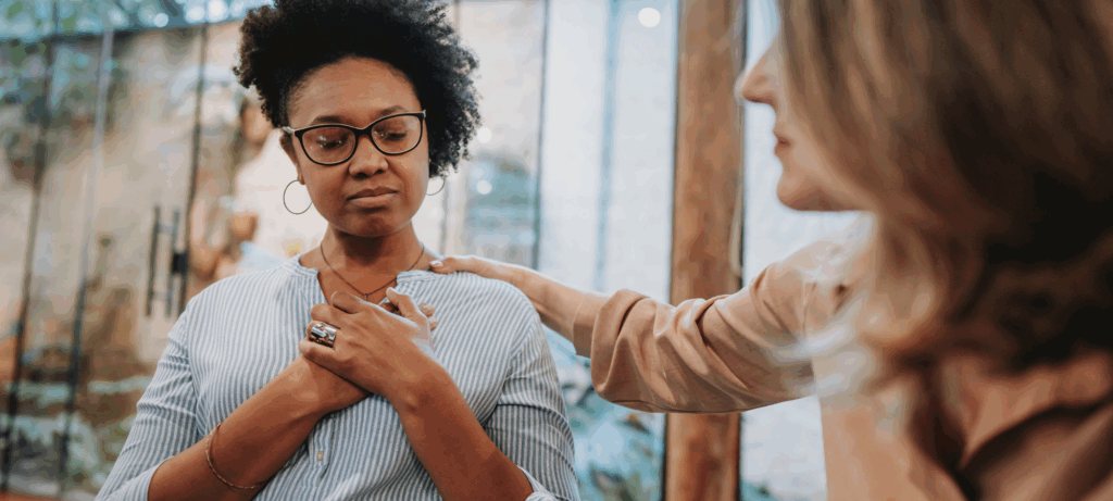 Black female with hands crossed across chest, eyes closed in relief behind glasses, being comforted by blonde woman (out of focus) in foreground