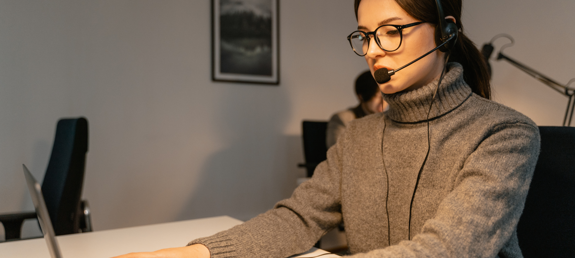 Brunette with low ponytail, glasses, and gray sweater wearing a headeset on a phone call in a dimly lit office